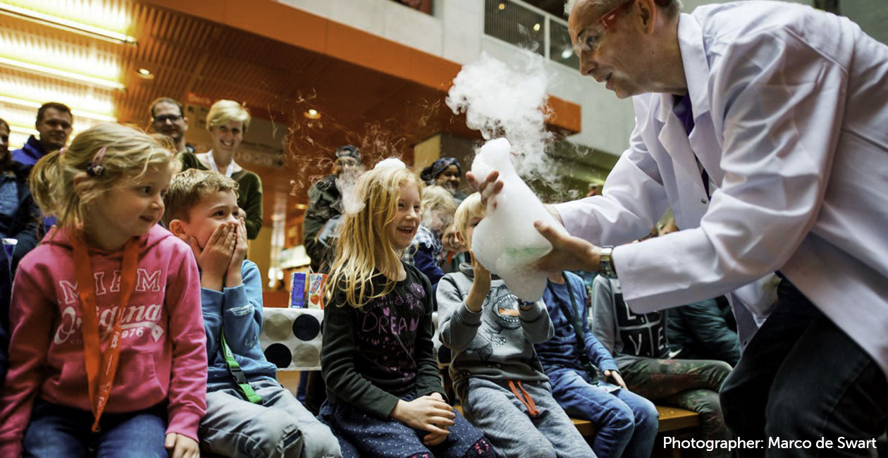 Mad scientist showing a beaker with smoke and bubble poring out to children while parents look on