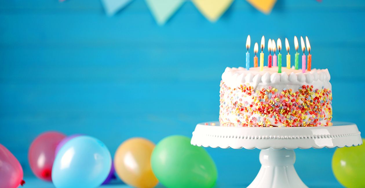 Delicious birthday cake on a white tray with burning candles and colorful balloons on table